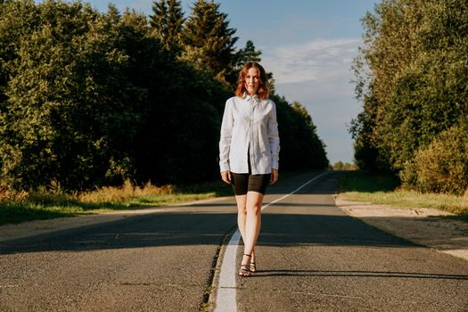 A red-haired woman in a white shirt walks along the road among the forest on a summer sunny day. A trip out of town.