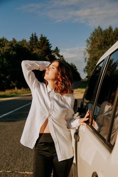A woman in a white shirt next to a white car on the road. A trip to nature, rest outside the city. Vertical photo