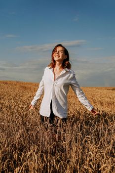 Happy young woman in a white shirt in a wheat field. Sunny day. Girl smiling, happiness concept. Hands to the side. Vertical photo