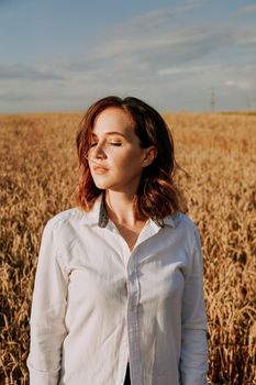 Portrait of a beautiful red-haired girl in a white shirt. She stands in a rye field on a sunny day. Calm facial expression. The concept of appeasement. Vertical photo