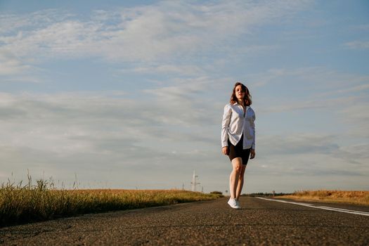 A red-haired woman in a white shirt walks along the road among the fields on a summer sunny day. A trip out of town