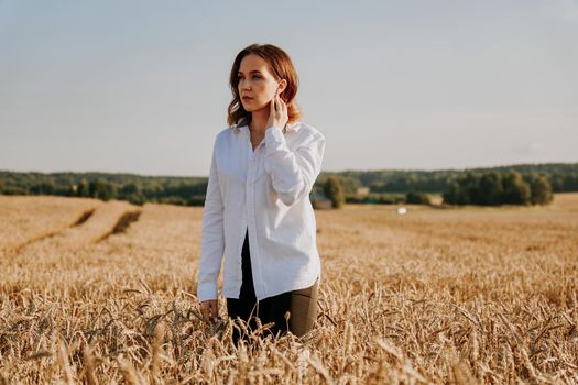 Portrait of a beautiful red-haired girl in a white shirt. She stands in a rye field on a sunny day. Calm facial expression. The concept of appeasement