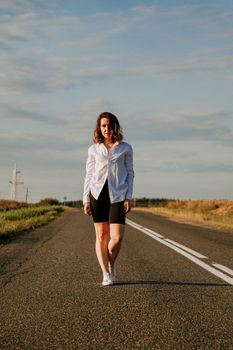 A red-haired woman in a white shirt walks along the road among the fields on a summer sunny day. A trip out of town. Vertical photo