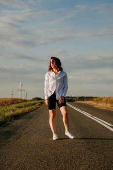 A red-haired woman in a white shirt walks along the road among the fields on a summer sunny day. A trip out of town. Vertical photo