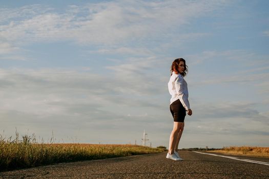 A red-haired woman in a white shirt walks along the road among the fields on a summer sunny day. A trip out of town