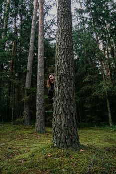 A young woman in a coniferous forest looks out from behind a tree. Happy young woman