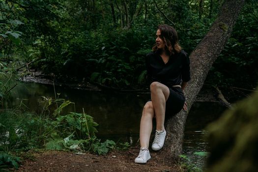 Woman in black clothes. Walk in a dark coniferous forest. Tracking and trip. Girl posing next to a tree above water