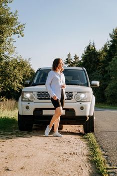 A woman in a white shirt next to a white car on the road. A trip to nature, rest outside the city. Vertical photo