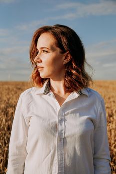 Portrait of a beautiful red-haired girl in a white shirt. She stands in a rye field on a sunny day. Calm facial expression. The concept of appeasement. Vertical photo