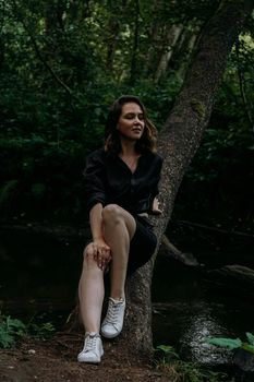 Woman in black clothes. Walk in a dark coniferous forest. Tracking and trip. Girl posing next to a tree above water