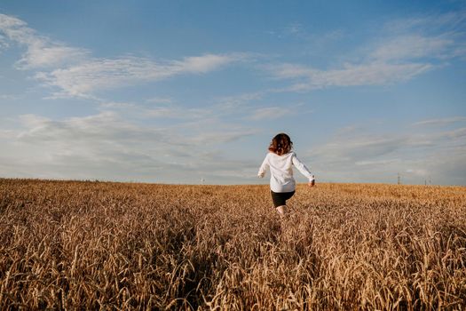 Happy woman in a white shirt runs in a wheat field on a summer day. Happiness and joy concept. Back view