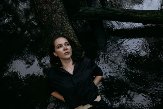 Woman in black clothes on a background of water, lies on a tree. Photo in the forest, the atmosphere of sadness and depression