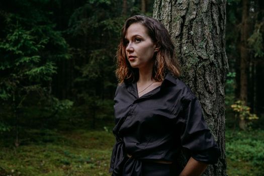 Young woman - close portrait in a dark pine forest. Woman in black shirt. She stands next to a tree