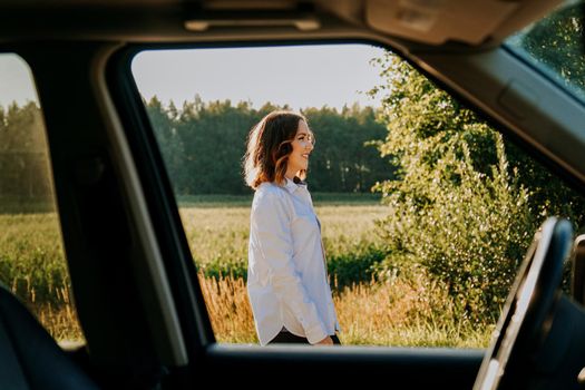 A beautiful young woman in a white shirt is resting outside the city. Outdoors near the forest and field. Photo through the car window. Car trip