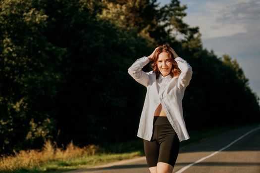 A red-haired woman in a white shirt walks along the road among the forest on a summer sunny day. A trip out of town.