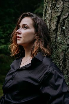 Young woman - close portrait in a dark pine forest. Woman in black shirt. She stands next to a tree