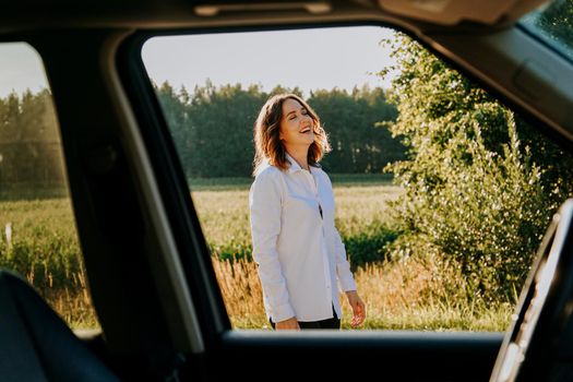 A beautiful young woman in a white shirt is resting outside the city. She is happy and laughs. Photo through the car window. Car trip