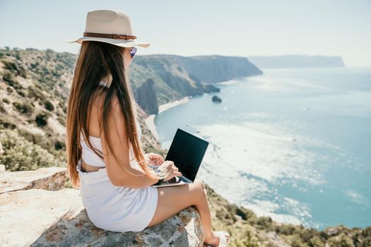 Successful business woman in yellow hat working on laptop by the sea. Pretty lady typing on computer at summer day outdoors. Freelance, travel and holidays concept.