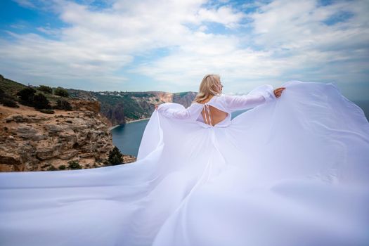 Blonde with long hair on a sunny seashore in a white flowing dress, rear view, silk fabric waving in the wind. Against the backdrop of the blue sky and mountains on the seashore