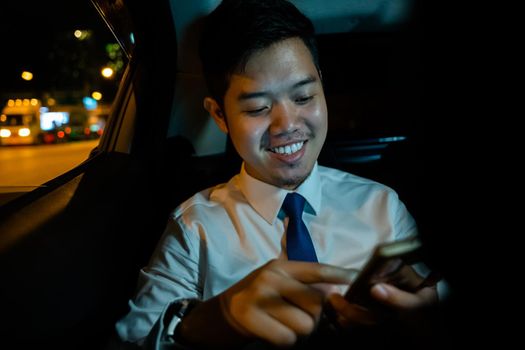 Handsome Asian young man in taxi work on late night sitting back seat and still work with mobile phone in city street, Businessman using smartphone in backseat of car near window at night