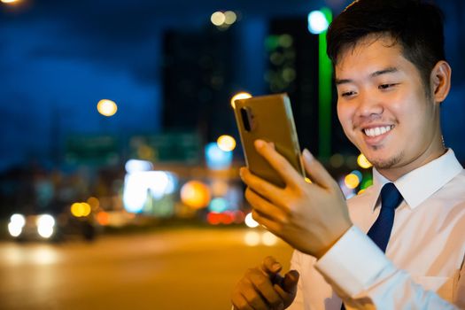 Portrait Asian businessman typing an sms message via smartphone after work near office at night city street, bokeh lights, young man walking and chatting with friends at social networks outdoor