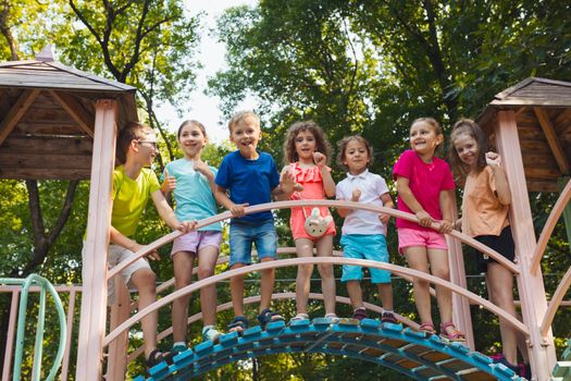 The elementary school children stand on a bridge in the playground and hold on to the railing. The kids dressed in colorful clothes are posing for the camera