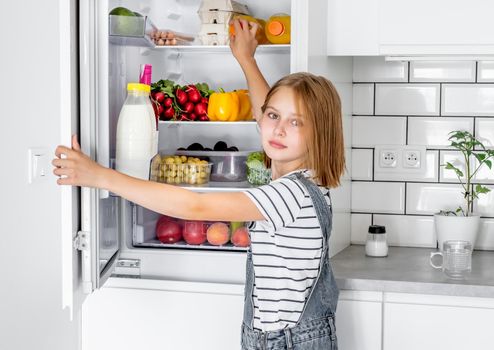 Preteen girl opens refrigerator with vegetables and fruits at kitchen. Female child kid and fridge with vegetarian vitamin diet