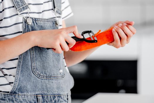 Preteen girl hand pilling carrot at kitchen closeup. Child with orange vegetables food cooking at home