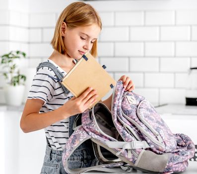 Preteen girl preparing backpack for school class lessons at home and put notebook inside. Female pupil with schoolbag
