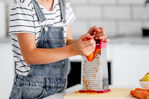 Girl child hands grinds carrot using grater at kitchen closeup. Pretty kid cooking vegetables at home