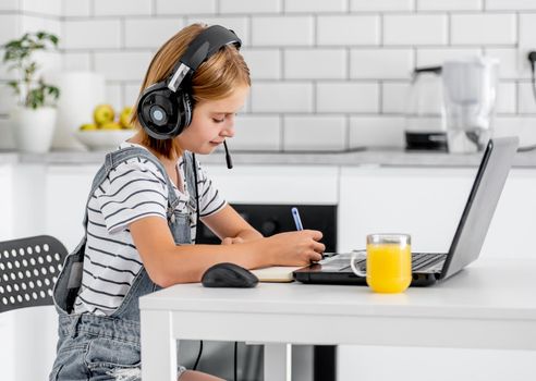 Preteen girl studying wearing headphones during online e-learning lesson at home at pandemic coronavirus. Child pupil making school video call with laptop and orange juice