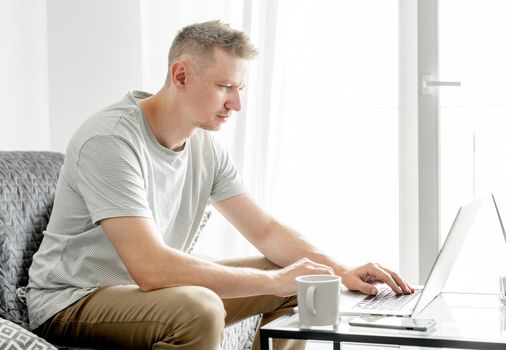 A young handsome man works at a laptop in a bright living room.