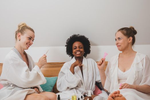 The young women in bathrobes are sitting on the bed and using skincare products
