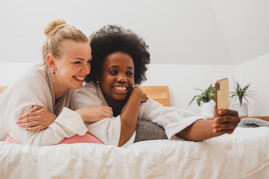 Two young women of different nationalities have a video chat at the spa. The women in bathrobes are lying on the bed and communicating to using a smartphone