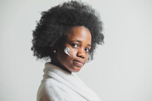 Portrait of a young woman in a bathrobe who is standing on a white background. The woman with a dab of cream on her face is looking at the camera