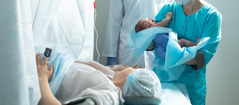 Nurse holds newborn baby close to mother in hospital, seconds after birth.
