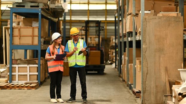 Full length of female managers and worker checking inventory in a warehouse with shelves full of cardboard boxes in the background.