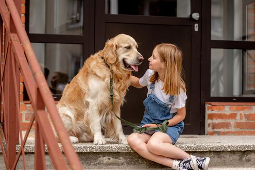 Preteen girl wearing jeans dress with golden retriever dog sitting outdoors in summertime. Pretty kid hugging purebred fluffy doggy pet in city