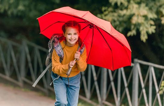 School girl with backpack and umbrella outdoors. Pretty child kid walking in rainy weather