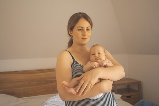 Mother holding baby sitting on the bed. New lifestyle with asleep newborn at bedroom