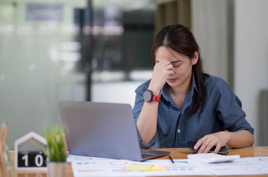 Asian women sitting in a home office With stress and eye strain.Tired businesswoman holding eyeglasses and massaging nose bridge. There are tablets, laptops, and coffee..