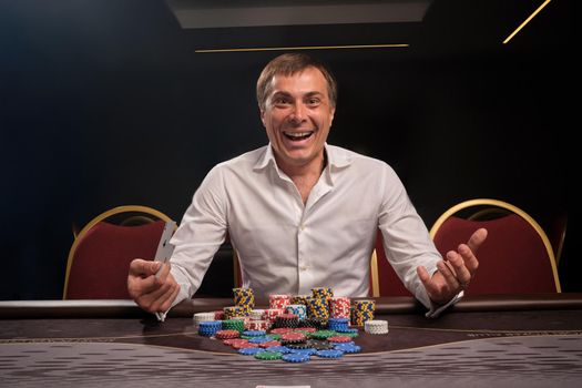 Young smiling man in a white classic shirt is playing poker sitting at the table in casino. He is rejoicing in winning and looking at the camera. Gambling for money. Games of fortune.