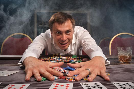 Young handsome man in a white classic shirt is playing poker sitting at the table in casino. He is rejoicing in winning holding his chips and looking at the camera. Gambling for money. Games of fortune. Smoke background.