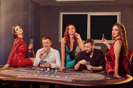 Group of a young players are posing at a poker table in casino and looking at the camera. Youth are making bets waiting for a big win. Gambling for money. Games of fortune.