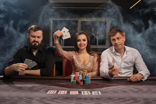 Cheerful friends are playing poker at casino in smoke. Girl has won and showing her cards, men had lost. Youth are making bets waiting for a huge win. Gambling for money. Games of fortune.