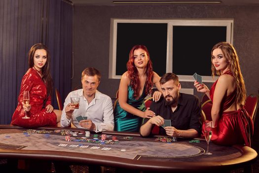 Group of a young smiling friends are playing at a poker table in casino and looking happy. Youth are making bets waiting for a big win. Gambling for money. Games of fortune.