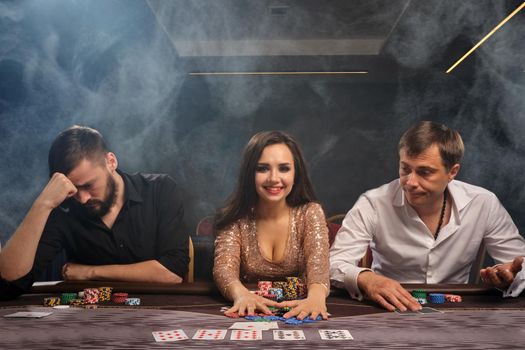 Young cheerful friends are playing poker at casino in smoke. Girl has won and guys had lost. Youth are making bets waiting for a huge win. Gambling for money. Games of fortune.