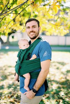 Dad with a baby in a sling standing under a tree. High quality photo