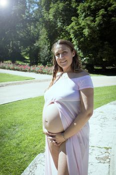 Seven month pregnant woman in pink transparent dress holding belly. Natural background