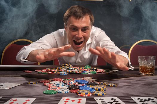 Young handsome guy in a white classic shirt is playing poker sitting at the table in casino. He is rejoicing in winning looking at his chips. Gambling for money. Games of fortune. Smoke background.